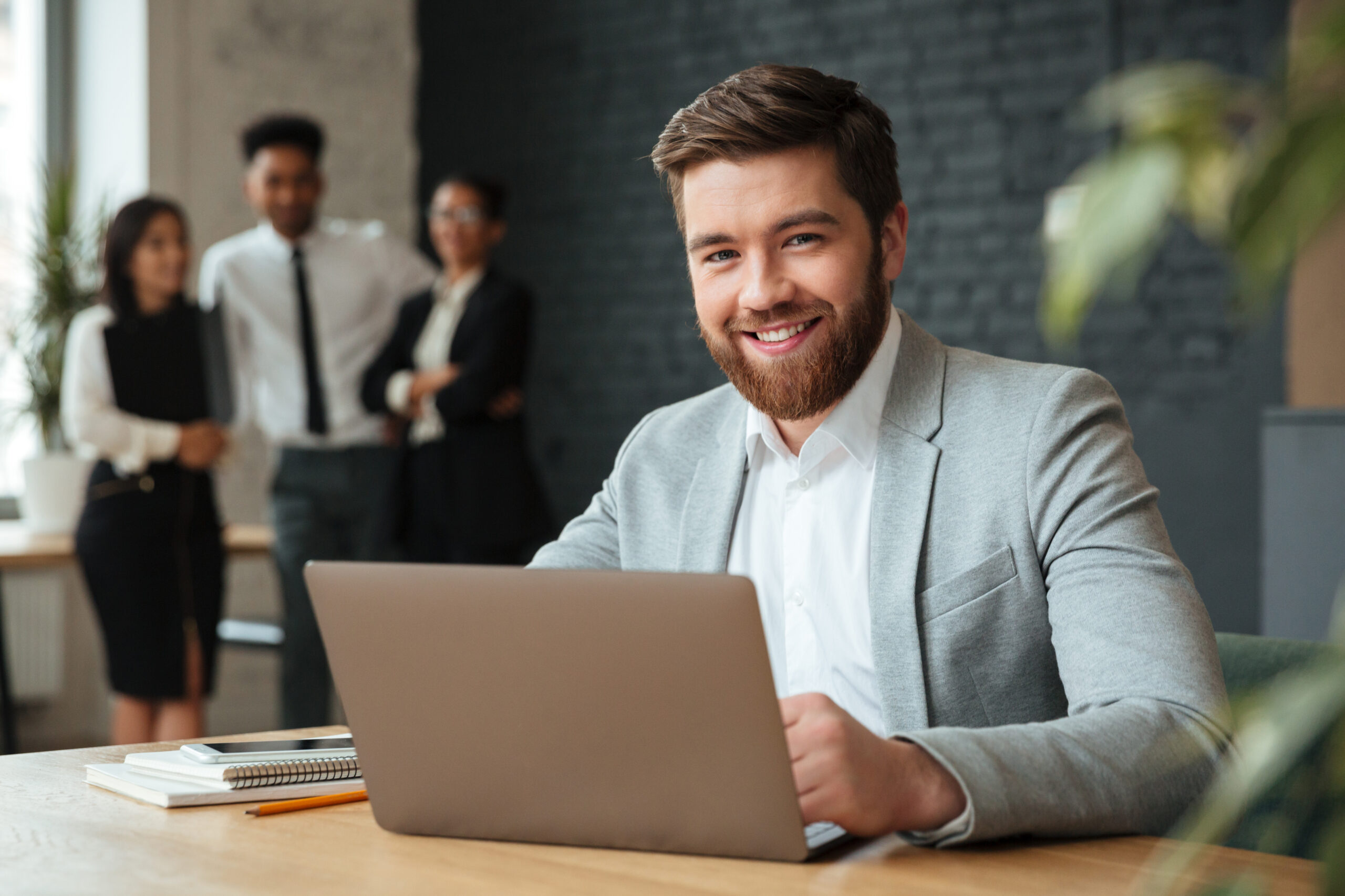 Image of cheerful young caucasian businessman sitting indoors using laptop computer. Looking camera.